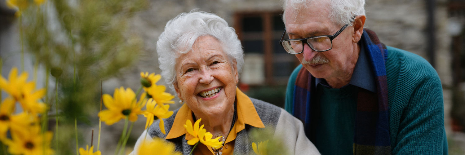 En ældre kvinde står bag nogle gule blomster og smiler. Bag hende står en mand og kigger på blomsterne.