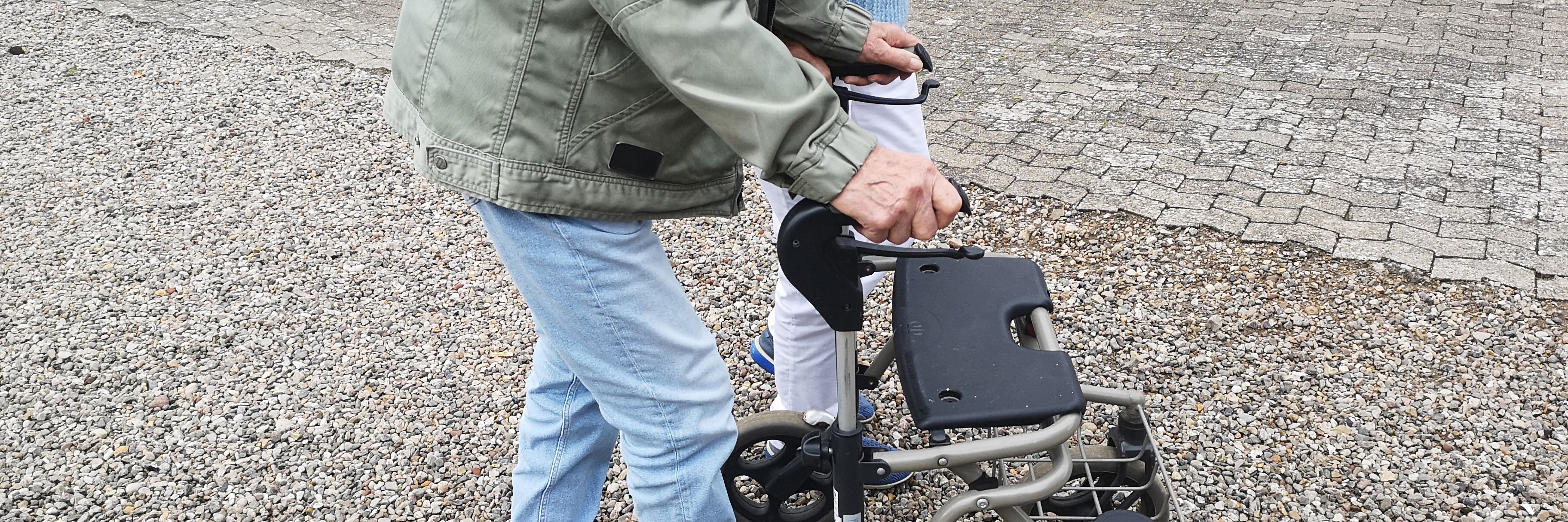 Et nærbillede af to personer, der går på en gårdsplads, hvoraf den ene går med rollator.